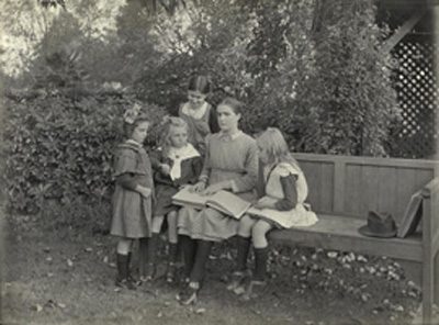 Tilly Aston sitting on a garden bench reading from a Braille book, surrounded by four young girls